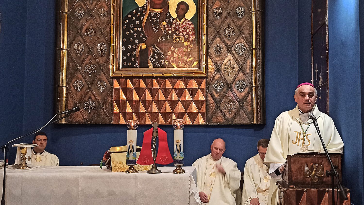 Biskup i kilku księży prowadzą nabożeństwo w kościele, stojąc i siedząc w pobliżu ołtarza. Za nimi znajduje się duża ikona Matki Boskiej z Dzieciątkiem, oprawiona w złote ramy na wzorzystym tle.