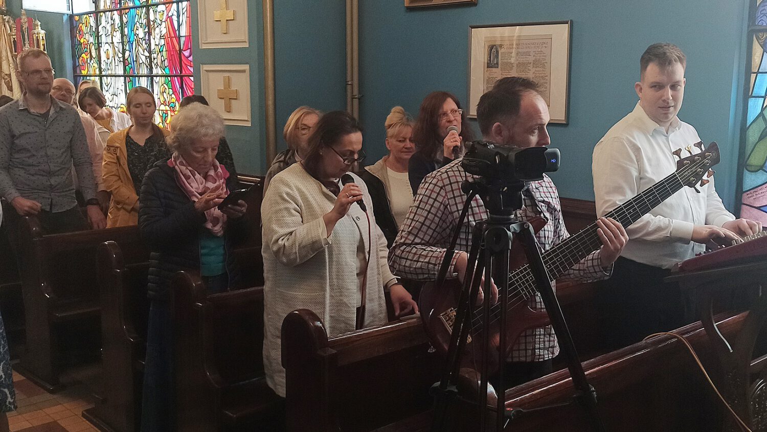 Grupa ludzi w kościelnej ławce śpiewa i trzyma mikrofony. Mężczyzna z gitarą i inny ze sprzętem wideo stoją z przodu. Kolorowe witraże i sztuka religijna zdobią ściany.