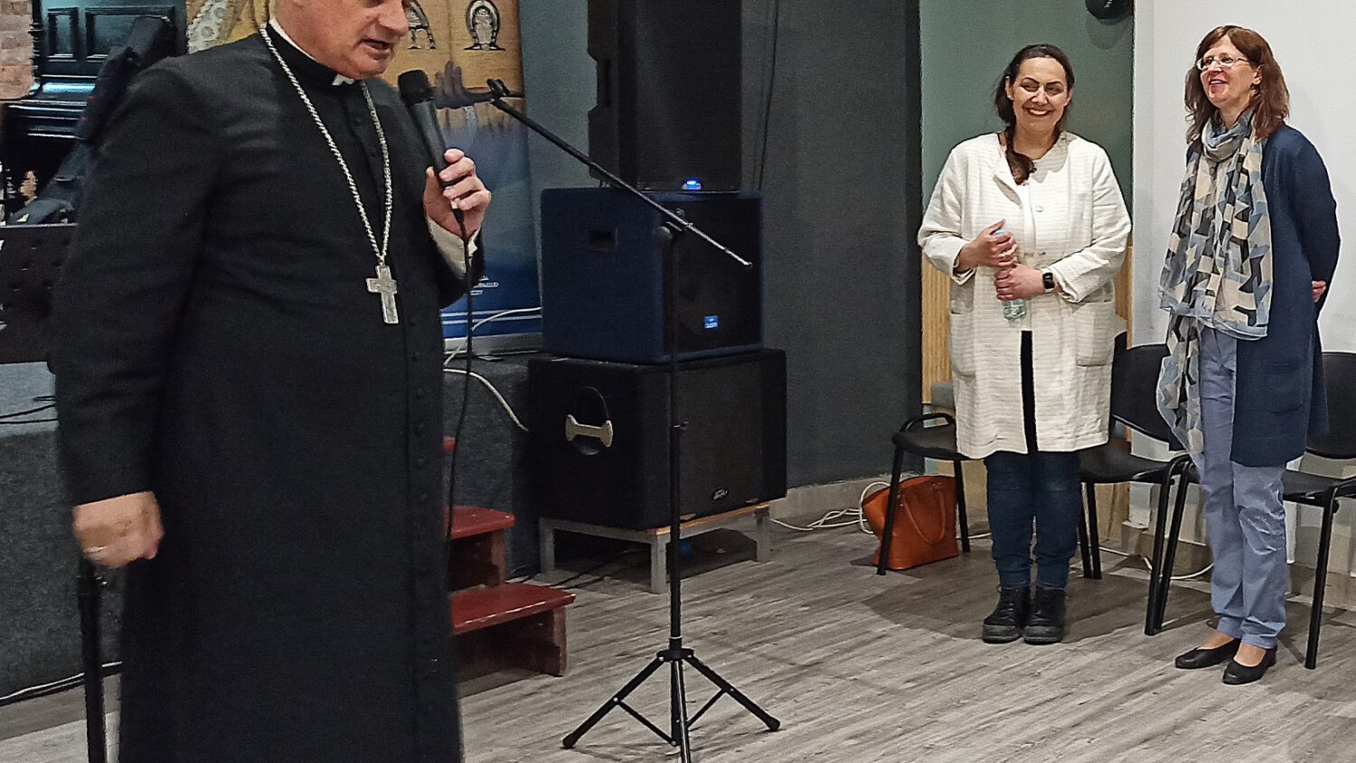 Biskup w czarnym stroju przemawia do mikrofonu, podczas gdy dwie kobiety stoją obok i uśmiechają się. W tle widoczny jest duży religijny obraz Jezusa. Scena wydaje się być w pomieszczeniu, prawdopodobnie podczas wydarzenia lub spotkania.