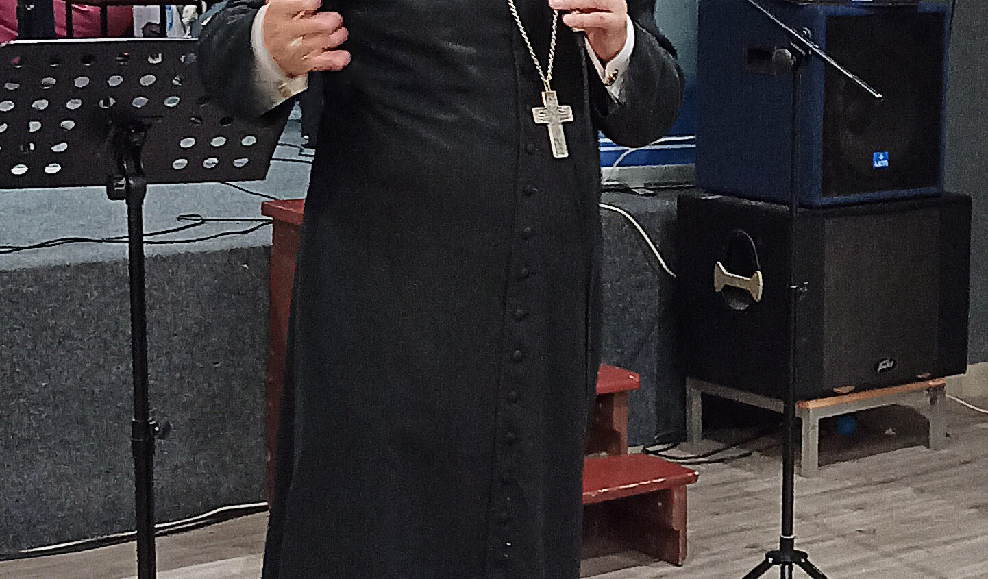 Biskup w czarnej szacie i różowym getrze przemawia do mikrofonu na scenie, gestykulując jedną ręką. Za nim znajduje się religijna ikona Jezusa i kilka statywów muzycznych.