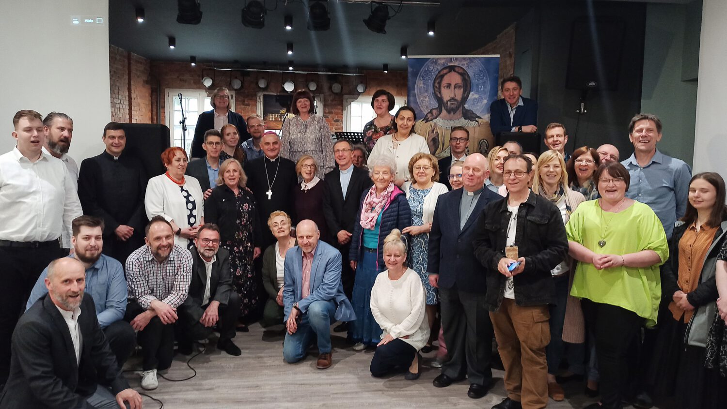 Duża grupa ludzi, mężczyzn i kobiet w różnym wieku, pozuje razem w pomieszczeniu przed mozaikowym wizerunkiem Jezusa. Niektórzy siedzą, a inni stoją, uśmiechając się do kamery w ciepło oświetlonym pomieszczeniu.