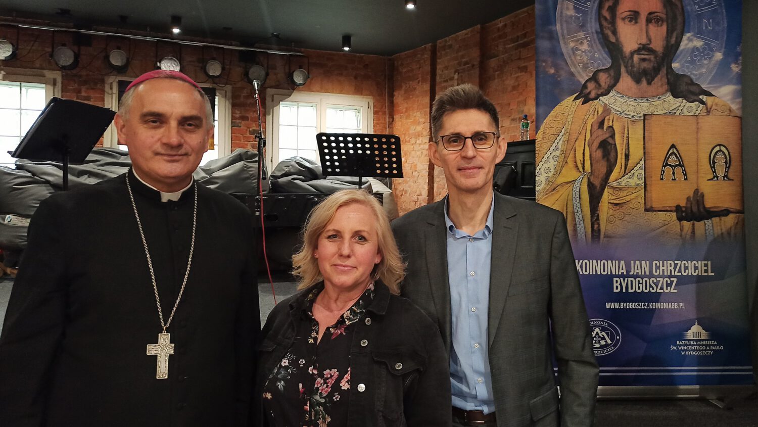 Trzy dorosłe osoby stoją w pomieszczeniu przed religijnym banerem przedstawiającym ikonę Jezusa. Mężczyzna po lewej stronie ma na sobie strój duchowny i krzyż, kobieta w środku ma na sobie czarną kurtkę, a mężczyzna po prawej stronie ma na sobie okulary i garnitur.