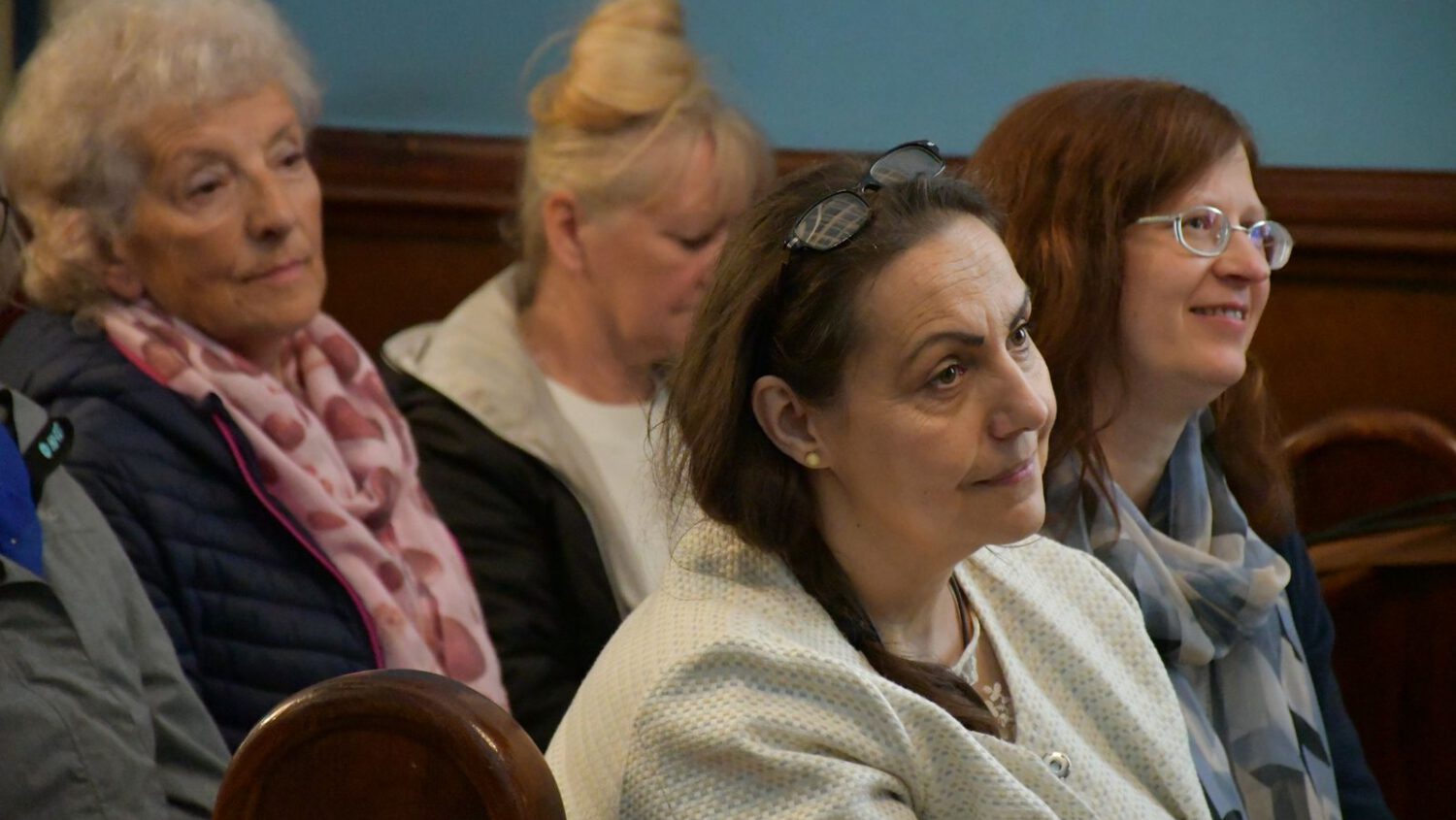 Cztery kobiety siedzą na drewnianych ławkach, uważnie słuchając. Mają na sobie swobodne ubrania i szaliki, a ich miny są neutralne lub zainteresowane. Tło jest niebieskie i brązowe, co sugeruje formalne lub społeczne otoczenie.