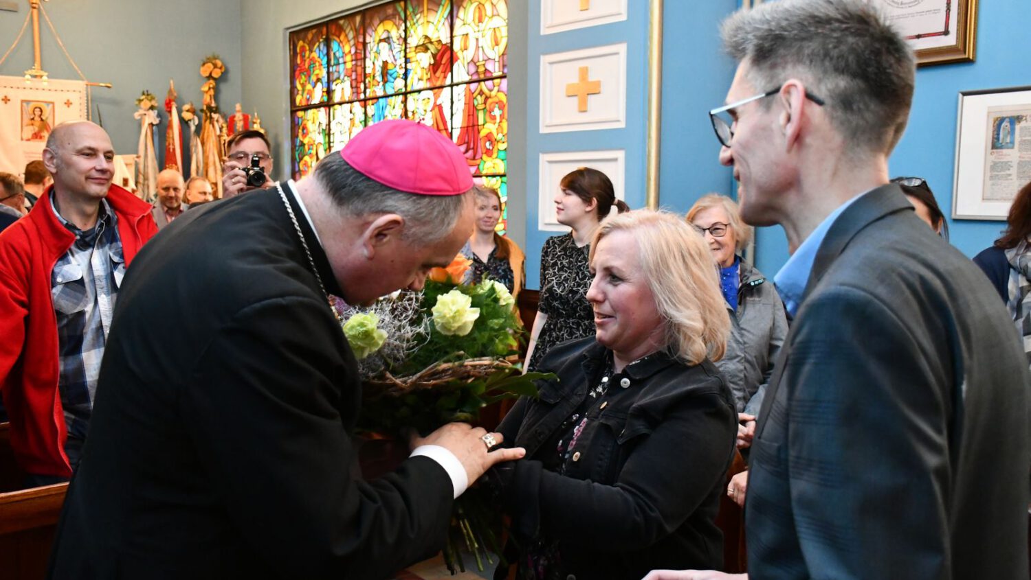 Biskup w różowej czapce kłania się, odbierając kwiaty od uśmiechniętej kobiety w kościele wypełnionym ludźmi. W tle widoczne są witraże i dekoracje religijne.