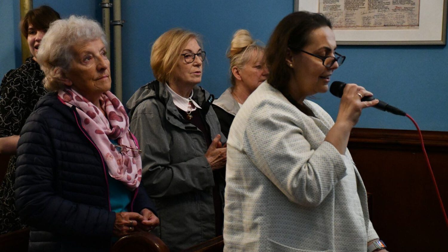 Kobieta w białej kurtce mówi do mikrofonu, podczas gdy cztery kobiety stoją za nią w pokoju z niebieskimi ścianami i oprawionym dokumentem na ścianie.