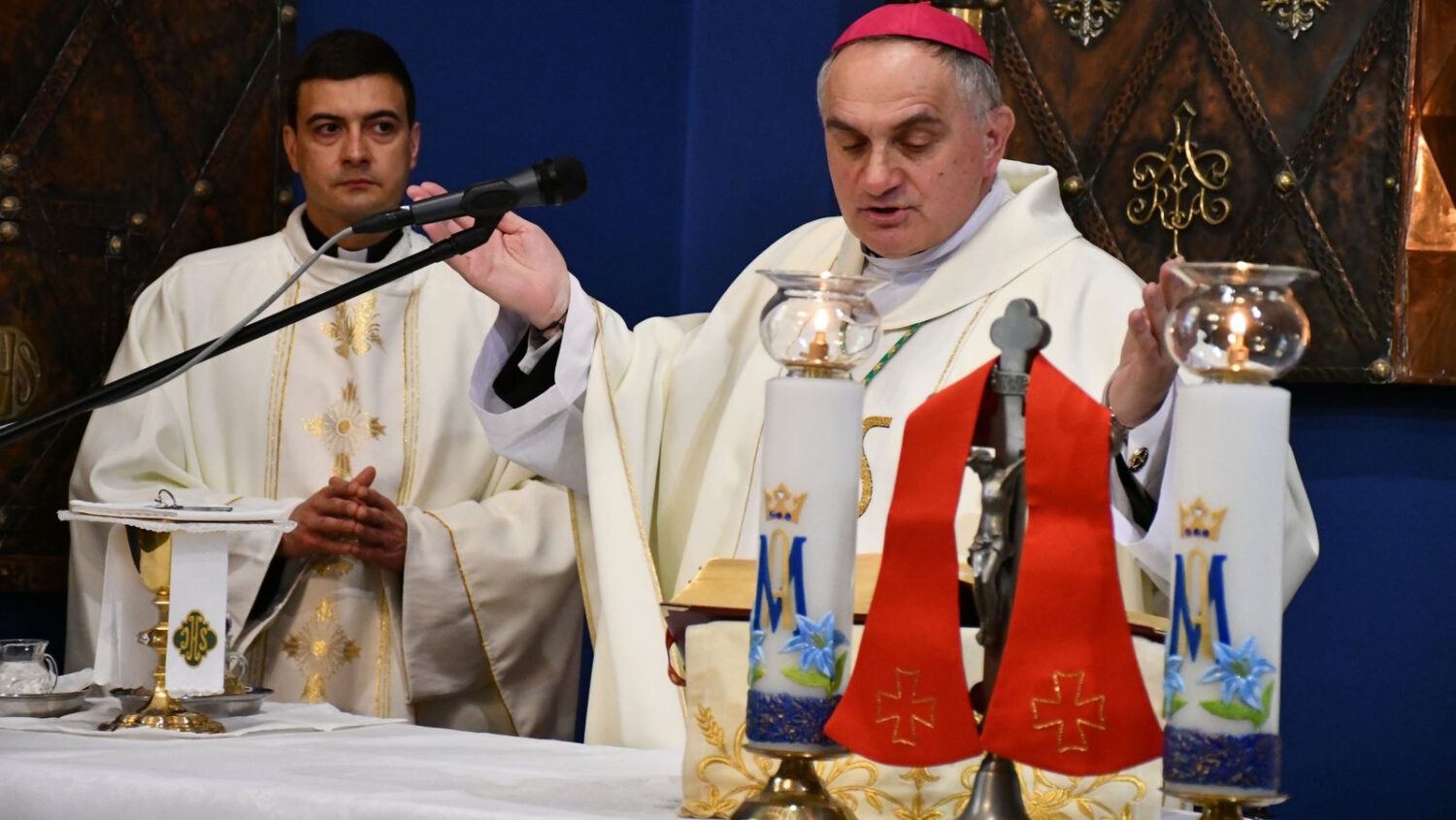 Biskup w białych szatach liturgicznych unosi ręce podczas odprawiania mszy przy ołtarzu, a obok niego stoi inny ksiądz. Ołtarz jest ozdobiony świecami, krucyfiksem i religijnymi dekoracjami.