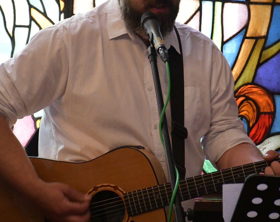 Mężczyzna z brodą i zaczesanymi do tyłu włosami gra na gitarze akustycznej i śpiewa do mikrofonu. Ma na sobie białą koszulę i stoi przed kolorowym witrażem.