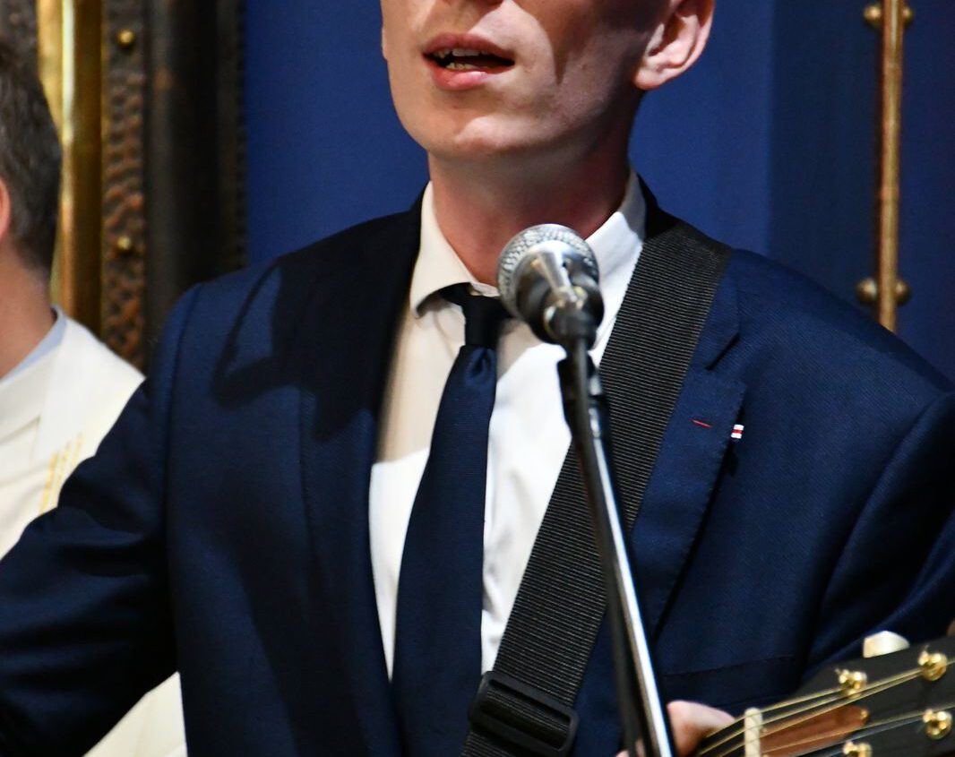Mężczyzna w ciemnym garniturze i okularach gra na gitarze akustycznej i śpiewa namiętnie do mikrofonu, z zamkniętymi oczami, z niebieskim tłem za sobą.